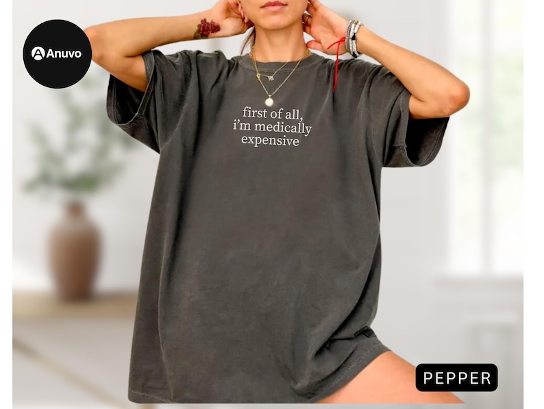 May include: A dark gray oversized t-shirt with the text "first of all, i'm medically expensive" in white. The shirt has short sleeves and a relaxed fit. The word "PEPPER" is in a black rectangle in the bottom right corner.
