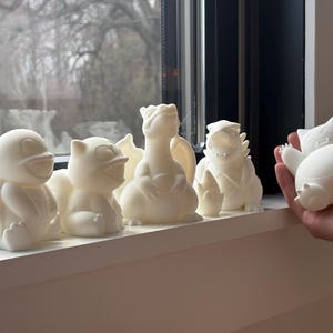 May include: A collection of five white 3D printed figures depicting various creatures, including a turtle, a small feline-like character, a winged dragon, a spiky-backed monster, and a round, sleeping character held in a hand.