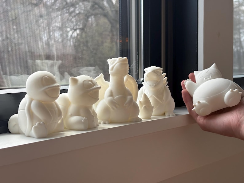 May include: A collection of five white 3D printed figures depicting various creatures, including a turtle, a small feline-like character, a winged dragon, a spiky-backed monster, and a round, sleeping character held in a hand.
