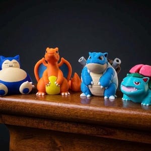 May include: Four colorful Pokémon figurines are displayed on a wooden surface. The collection includes a blue Snorlax, an orange Charizard, a blue Blastoise, and a green Venusaur with a pink flower. The figures are detailed and stand out against the dark background.