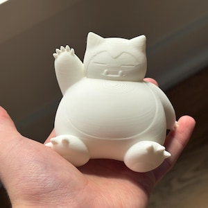 May include: A white, three-dimensional printed figurine of a round, seated creature with pointed ears, closed eyes, and two small fangs. The creature is depicted with one arm raised in a waving gesture, featuring visible horizontal layer lines from the printing process.
