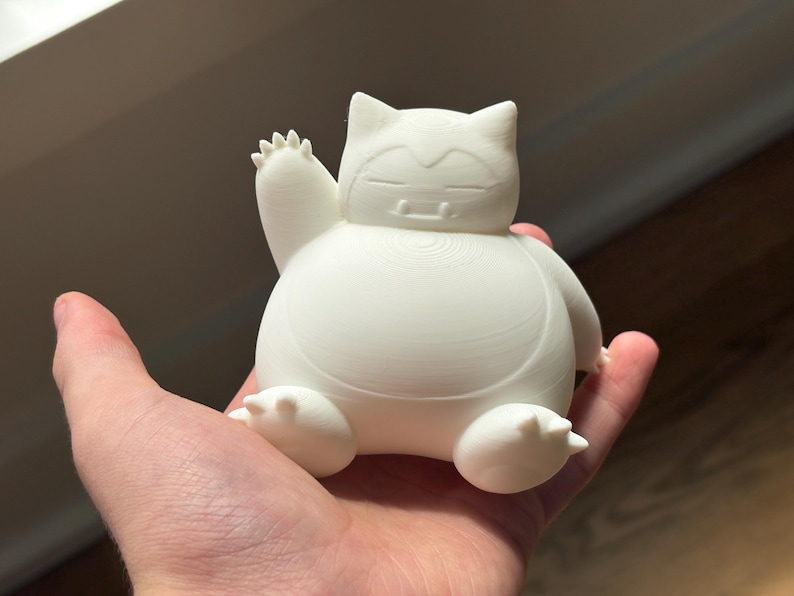 May include: A white, three-dimensional printed figurine of a round, seated creature with pointed ears, closed eyes, and two small fangs. The creature is depicted with one arm raised in a waving gesture, featuring visible horizontal layer lines from the printing process.