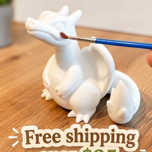May include: A white, unpainted ceramic dragon figurine with a smooth matte finish, featuring a rounded body, small wings, and a flame-tipped tail. A thin paintbrush with a blue handle is positioned near the dragon's snout for painting.
