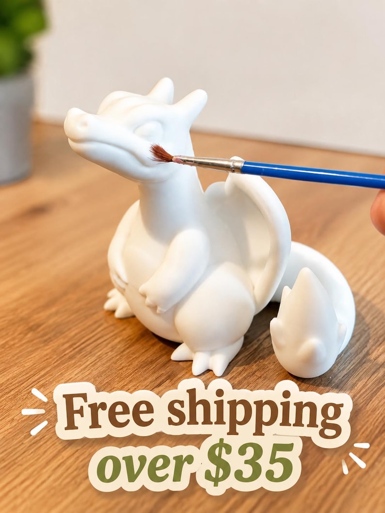 May include: A white, unpainted ceramic dragon figurine with a smooth matte finish, featuring a rounded body, small wings, and a flame-tipped tail. A thin paintbrush with a blue handle is positioned near the dragon's snout for painting.