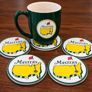 Op de afbeelding: Een groene koffiemok en onderzetters set. De mok en onderzetters tonen het Masters-logo: een gele omtrek van een golfbaan met een rode vlag en het woord "Masters". De onderzetters zijn rond met een witte rand.