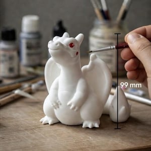May include: A white dragon-like creature figurine with red eyes and red cheek spots. A vertical line indicates a height of 99 mm. The text at the bottom reads: Size varies slightly with type of Pok&eacute;mon. The figurine has wings and a tail.