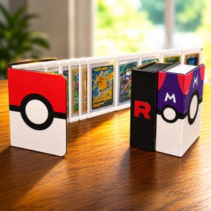May include: A collection of trading card display cases featuring a red and white Poké Ball design, a black case with a red letter R, and a purple and white Master Ball design. The cases are arranged to hold multiple individual trading cards.