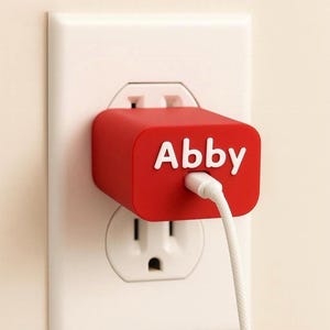 以下が含まれることがあります： 白い壁のコンセントに、白い文字で「Abby」と書かれた赤い充電アダプターが差し込まれています。白い充電ケーブルがアダプターに接続されています。コンセントは白い壁に取り付けられています。