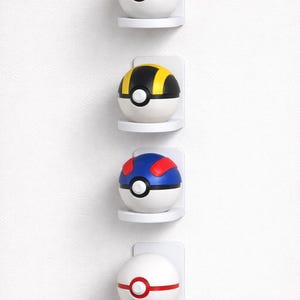 Könnte beinhalten: Vier farbenfrohe Pokéball-Repliken, die auf einzelnen weißen Regalen vor einer weißen Wand ausgestellt sind. Die Bälle sind rot, gelb, blau und weiß mit schwarzen und weißen Akzenten. Jeder Ball hat einen kreisförmigen Knopf.