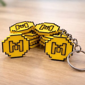 Può includere: Una collezione di portachiavi a forma di moneta gialla pixelata con contorno nero e portachiavi argentato. Ogni moneta presenta un design "M" nero. I portachiavi sono impilati su una superficie di legno chiaro.