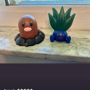 May include: Two small figurines, one brown, cylindrical creature with a pink nose and black eyes, and one blue, round creature with green, leaf-like protrusions on top, red eyes, and a small open mouth, both displayed as decorative items.