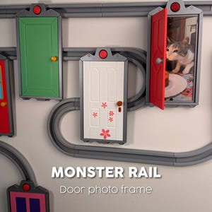 May include: A wall-mounted display featuring gray plastic tracks connecting multiple rectangular door-shaped frames. The frames are painted in various colors including green, white with pink flower decals, and red, which holds a photo of a cat.