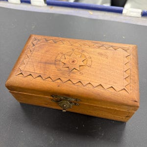 Puede incluir: Pequeña caja rectangular de madera clara. La tapa presenta un diseño de estrella tallada y bordes en zigzag. Tiene un cierre de latón. Ideal para guardar joyas u objetos pequeños.