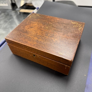 May include: A rectangular wooden box with a dark brown finish. The box has a small keyhole on the front and a decorative design on the top. The wood grain is visible, and the box appears to be vintage or antique.
