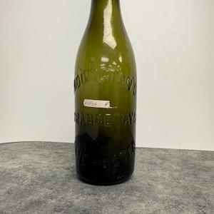 May include: An antique green glass bottle with a black cap. The bottle has embossed text that reads "ADDINGTON'S" and "FRANCE WAYS" and "WINCHESTER". A small white tag with the number "#9864" is attached.