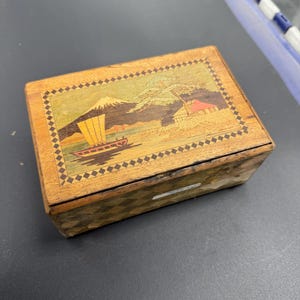 May include: A rectangular wooden box with a decorative inlaid design. The lid features a landscape scene with a mountain, boat, and trees. The box has a diamond pattern border and is made of light brown wood.