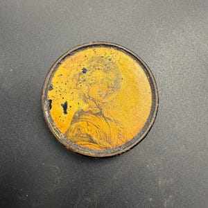 May include: A circular antique medallion featuring a portrait. The portrait is rendered in yellow and brown tones, set within a dark brown border. The medallion has a distressed, aged appearance.