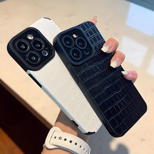May include: Two phone cases with a crocodile skin texture. One case is white, and the other is black. Both cases have a black camera lens protector. The cases are designed to protect the phone from scratches and damage.