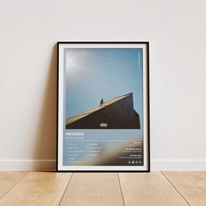 May include: Framed poster featuring the album "Freudian" by Daniel Caesar. The artwork shows a person on a building against a blue sky. The poster includes a tracklist and album details in a black frame, displayed on a wooden floor.