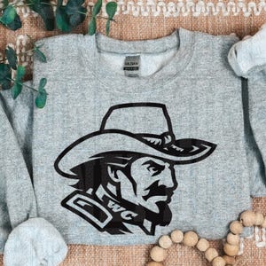 May include: A light gray sweatshirt featuring a black graphic of a cowboy wearing a hat. The graphic includes the letters "WC". The sweatshirt is made of a soft material.