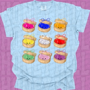 Puede incluir: Camiseta azul claro con una cuadrícula de nueve cajas de regalo de dibujos animados. Cada caja contiene un artículo de diferente color y tiene un lazo en la parte superior. Los colores incluyen rosa, azul, amarillo, blanco, rojo, verde, rosa y morado.