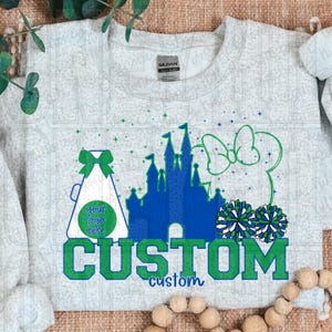 Peut inclure: Un sweat-shirt gris clair avec un motif personnalisé représentant un château bleu, un nœud vert et des pompons. Le mot "CUSTOM" est en lettres vertes, avec "custom" en plus petit en dessous. Un mégaphone de pom-pom girl est également présent.