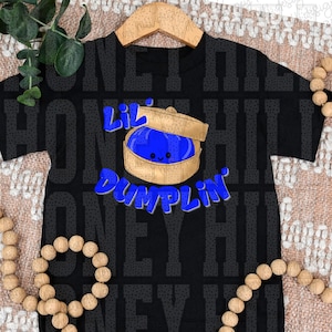 Puede incluir: Camiseta negra con un gráfico de una bola de masa hervida azul en una vaporera de bambú. El texto "Lil Dumplin" es azul, con un estilo de dibujos animados. La camiseta se muestra en una percha de madera.