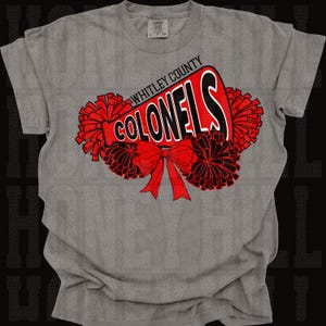 May include: A grey t-shirt with a red and black design. The design features the text "WHITLEY COUNTY COLONELS" in a megaphone shape, with red pom-poms and a red bow. The shirt has a crew neck and short sleeves.