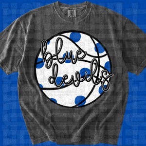 Peut inclure: Un t-shirt gris anthracite à manches courtes avec un grand motif circulaire représentant un ballon de basket blanc à pois bleus. Le texte "blue devils" est écrit en lettres cursives noires au centre du motif du ballon.