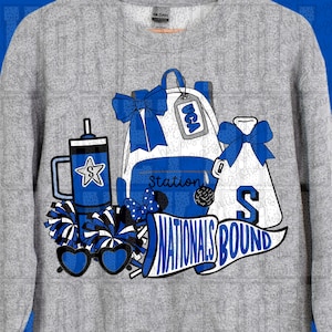 May include: A gray sweatshirt featuring a graphic design with blue and white elements. The design includes a backpack, pom-poms, sunglasses, and the text "NATIONALS BOUND". A blue tumbler with a star is also visible.