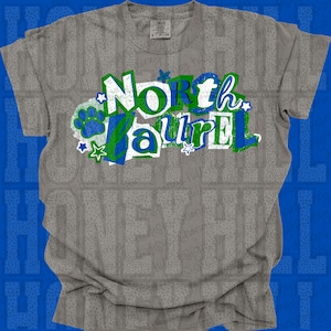 May include: A gray t-shirt with the words "North Laurel" in a layered design of white, green, and blue. The design includes a paw print and stars. The shirt is made of a soft material.