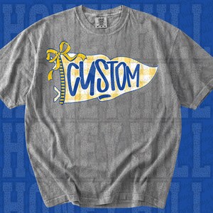 May include: A grey short-sleeve t-shirt featuring a graphic of a yellow and white checkered pennant flag with a yellow bow at the left side. The word CUSTOM is printed in bold blue script across the centre of the pennant flag design.
