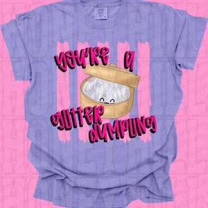 Puede incluir: Camiseta lila con la frase "You're a Glitter Dumpling". El diseño presenta una caricatura de una empanadilla en una vaporera de bambú, con rayas verticales rosas de fondo.
