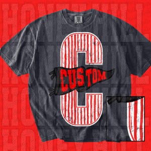 May include: A charcoal gray short-sleeve t-shirt featuring a large, distressed white and red vertical striped letter C graphic. Overlaid across the center of the letter is a black pennant flag shape containing the red text CUSTOM.