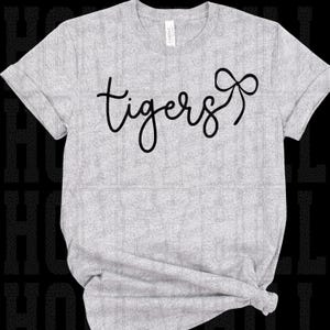 Könnte beinhalten: Hellgraues T-Shirt mit dem Wort "tigers" in schwarzer Schrift und einer Schleifengrafik. Das Shirt besteht aus einem weichen Material und hat kurze Ärmel. Das Shirt ist am unteren Ende geknotet.