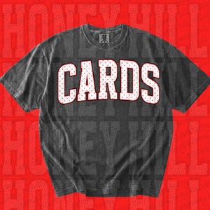 May include: A charcoal grey short-sleeve t-shirt featuring the word CARDS printed in large, white, block-style letters with a red polka dot pattern. The shirt has a crew neckline and a visible brand tag on the inside of the collar area.