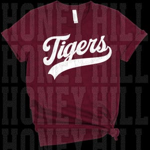 May include: A maroon short-sleeved t-shirt featuring the word Tigers in white script lettering across the chest. The text is written in a bold, cursive font with a decorative underline swoosh. The shirt has a crew neckline and a standard fit.