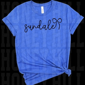 Könnte beinhalten: Blaues T-Shirt mit dem Wort "sundale" in schwarzer Schrift. Die Ärmel sind hochgekrempelt und das T-Shirt ist am Saum geknotet. Das Wort "sundale" hat am Ende eine Schleife.