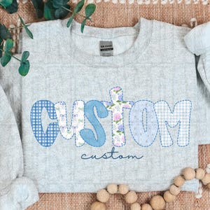 May include: Light gray sweatshirt with the word "custom" in a patchwork design. The letters are in blue gingham, floral, and solid blue patterns. The word "custom" is also written in script below the main text.