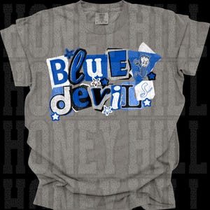 Puede incluir: Camiseta gris con un diseño gráfico que dice "Blue Devil" en letras de bloque azules y blancas. El diseño incluye un pequeño diablo de dibujos animados y formas de estrella. La camiseta está hecha de un material suave.