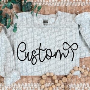 May include: A light gray sweatshirt with the word "Custom" in a black script font. The sweatshirt has long sleeves and a crew neck. The background has a subtle pattern and is decorated with greenery and wooden beads.