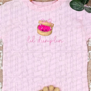 Puede incluir: Camiseta rosa claro con una ilustración de dibujos animados de una bola de masa rosa dentro de una caja marrón con un lazo rosa. El texto "lil dumplin" está escrito en rosa debajo.