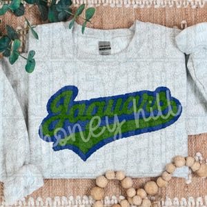 May include: A light gray sweatshirt with the words "Honey Hill" in a blue and green design. The text is in a stylized font and is centered on the front of the shirt. The sweatshirt has a crew neck and long sleeves.