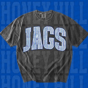 Puede incluir: Una camiseta de manga corta en color gris carbón con la palabra JAGS impresa en letras grandes y llamativas de color azul con un patrón de rayas verticales blancas. La tela tiene una textura lavada y el cuello incluye una pequeña etiqueta negra.