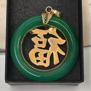 May include: A green jade pendant with a gold-colored Chinese character in the center. The pendant is circular with a gold-colored rope-like border. The pendant is displayed in a black box.