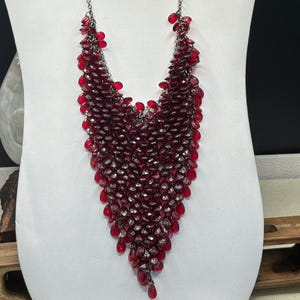 Op de afbeelding: Een lange, donkerzilveren ketting met een waterval van dieprode kralen. De kralen zijn samengeklonterd en vormen een V-vorm die over de borst hangt. De ketting wordt getoond op een witte mannequin.