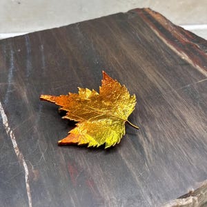 May include: A metallic maple leaf brooch featuring a gradient color scheme that transitions from deep orange and red at the edges to a bright, vibrant yellow in the center, showcasing a detailed, textured surface that mimics natural leaf veins.