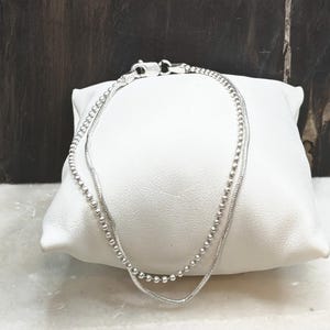 May include: A double-strand silver bracelet featuring one delicate ball chain and one smooth snake chain. The two chains are connected by a single lobster claw clasp, creating a layered look with a polished metallic finish and a bright silver tone.