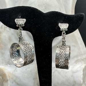 May include: Silver-toned dangle earrings with intricate openwork designs. Each earring features a decorative top piece connected to a large, cylindrical hoop with detailed patterns. The earrings are displayed on a black stand against a white and black background.