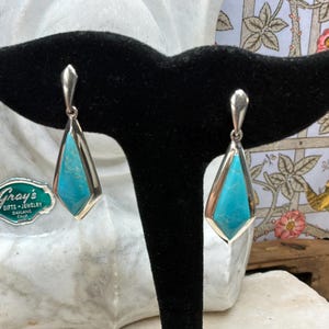 May include: A pair of turquoise and silver earrings on a black display stand. The earrings have a geometric design with a turquoise stone set in a silver frame. The earrings are displayed in front of a marble bust and a floral patterned background.